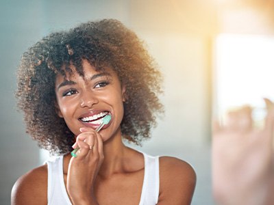 A young, smiling woman brushing her teeth in front of a bathroom mirror